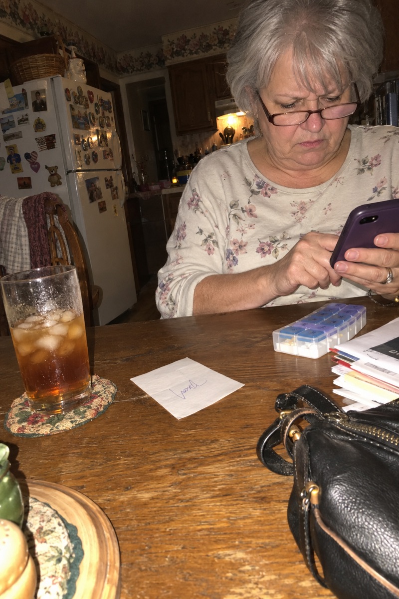 A woman in her sixties at her kitchen table at night, reading on her phone next to a pill organizer